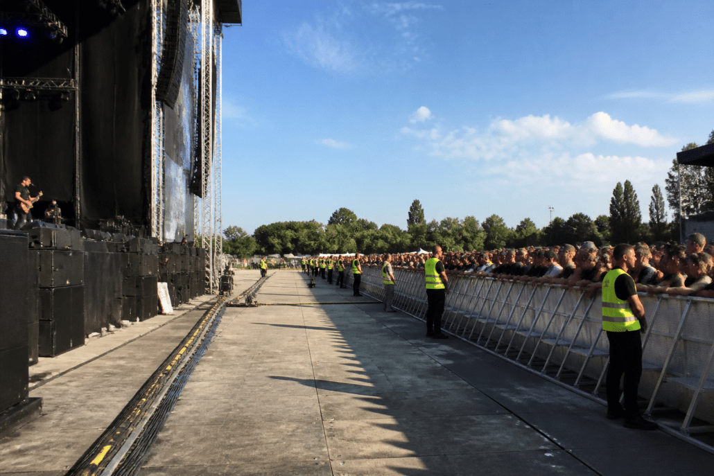 Concierto al aire libre con vallas y franja de seguridad entre escenario y público, personal de seguridad visible.