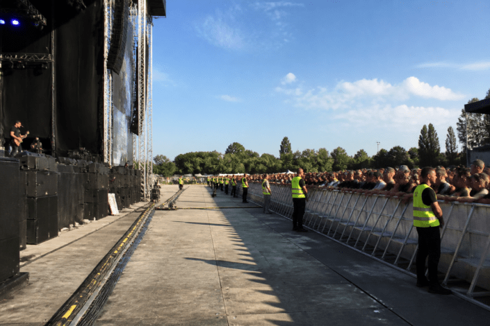 Concierto al aire libre con vallas y franja de seguridad entre escenario y público, personal de seguridad visible.